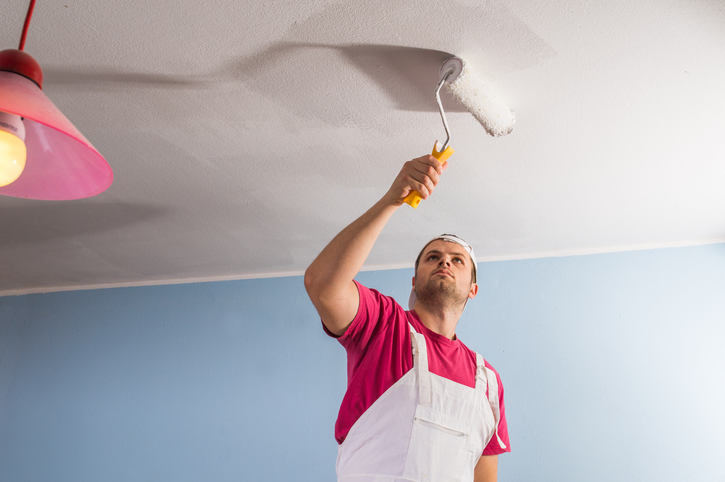 Man painting the ceiling of a living room white. Attempting to hake his low ceiling appear higher. 